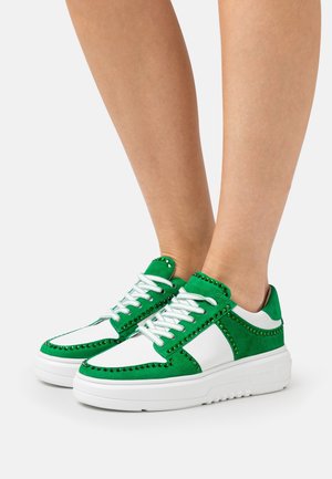 Jambes portant des baskets vertes et blanches avec des lacets blancs et des clous verts sur des panneaux en daim, debout sur un fond blanc uni.