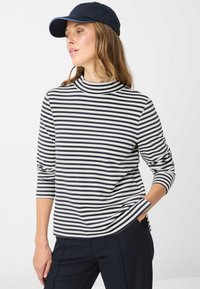 Gestreiftes Langarm-Oberteil in Weiß und Marineblau mit hohem Halsausschnitt und lockerem Schnitt. Kombiniert mit marineblauen Hosen und passender Mütze.