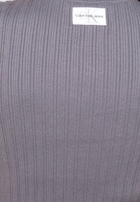 Szary, prążkowany top z dzianiny z białą prostokątną metką z napisem "Calvin Klein Jeans", z pionowym wzorem i dopasowanym krojem.