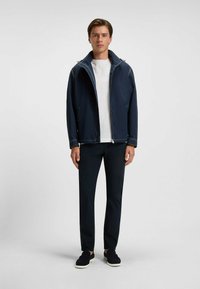 Marineblauwe jas met een hoge kraag en lichte stiksels, gecombineerd met een witte t-shirt, donkere broek en zwarte instapschoenen.