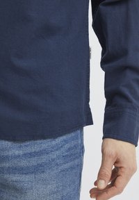 Diepblauwe langekiefshirt met een zachte textuur, gebogen zoom en zichtbare zijsplitten. De mouwen hebben een manchetdetail. Draagt het met lichtblauwe jeans.