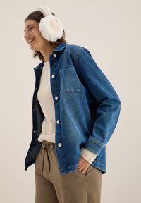 Camicia in denim di blu medio con tasca, bottoni bianchi e vestibilità rilassata. Indossata sopra un maglione leggero a maglia e pantaloni beige. Cuffiette in pelliccia bianca.