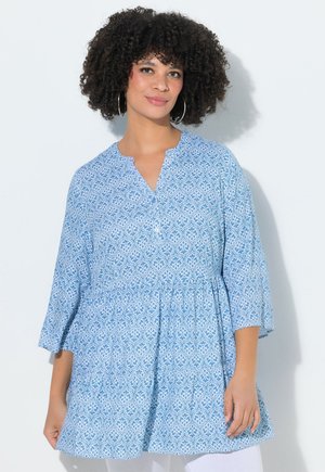 Donna con capelli ricci che indossa grandi orecchini a cerchio e una tunica blu con motivi e maniche a tre quarti, su sfondo chiaro.