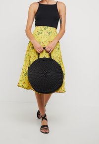 Cirkulär svart vävd handväska med en rund botten och två handtag, matchad med en gul blommig kjol och en svart figurnära topp.