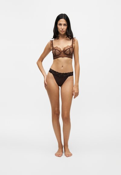 Femme debout pieds nus portant un soutien-gorge en dentelle marron et des sous-vêtements assortis bordés de dentelle sur un fond blanc uni.