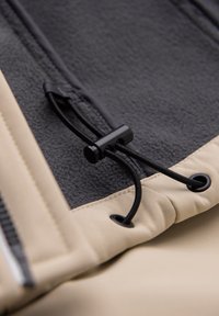 Cordon élastique noir avec bouton de réglage sur tissu beige et gris foncé, fixé par des œillets métalliques sur un vêtement.