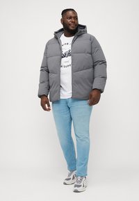 Doudoune grise avec capuche, présentant des sections matelassées, portée sur un t-shirt graphique blanc. Assortie avec un jean bleu clair et des baskets grises.