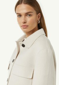 Crème kleurige wollen mix jas met een kraag, zwarte knopen en een opgestikte zak op de borst, getoond op een model tegen een neutrale achtergrond.