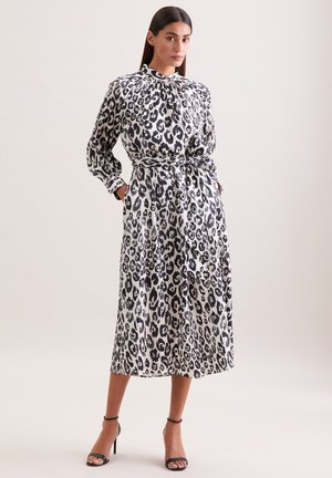 Kobieta stojąca z rękami w kieszeniach, ubrana w czarno-białą midi sukienkę w cętki lamparta i czarne sandały na wysokim obcasie.