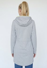 Žena s blond koňským chvostom, oblečená v dlhom svetlosivom kabáte s kapucňou, zobrazená zozadu na jednoliatom bielom pozadí.