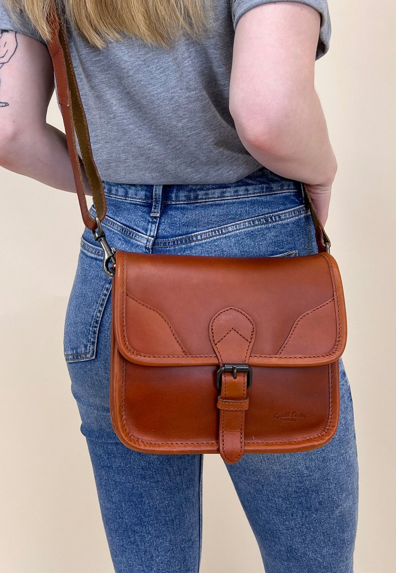 Hnědá kožená crossbody taška s klopou, kovovou sponou a zakřivenými detaily. Nositelná na těle spolu s džínami a šedou košilí.