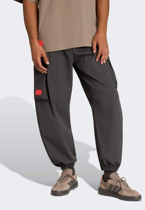 Person trägt dunkelgraue Cargohose mit elastischen Bündchen und beigefarbene Turnschuhe mit schwarzen Streifen, steht vor einem schlichten Hintergrund.