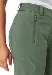 Grüne Outdoor-Hosen aus leichtem Stoff, mit einer Reißverschlusstasche, texturiertem Bund und einem eingeprägten Logo am Oberschenkel.