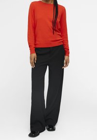 Suéter de punto rojo con mangas largas combinado con pantalones de pierna ancha negros de talle alto; unos mocasines negros brillantes completan el look.