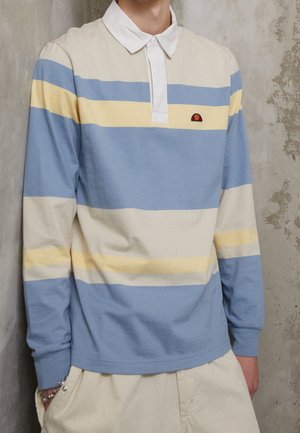 Personne portant un polo à manches longues avec des rayures horizontales bleu clair, crème et jaune et un pantalon beige, debout contre un mur texturé.