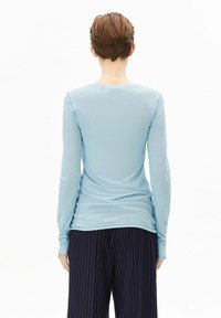 Hellblaues Langarmoberteil mit rundem Halsausschnitt und glatter Textur, kombiniert mit dunklen, gestreiften Hosen. Ansicht von hinten.