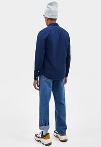 Granatowa koszula na guziki z lekko zaokrąglonym dołem, w zestawieniu z jasnoniebieskimi dżinsami. Całość dopełnia szara czapka i wielokolorowe tenisówki.