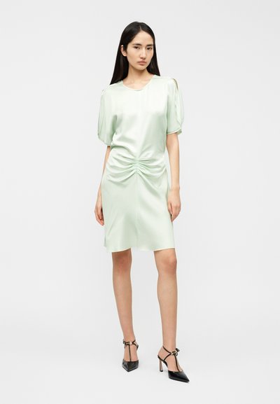Φόρεμα από σατέν ανοιχτό πράσινο με κοντά φουσκωτά μανίκια, πιέτες μπροστά και μήκος μέχρι το γόνατο. Το μοντέλο φοράει μαύρα ψηλοτάκουνα παπούτσια.