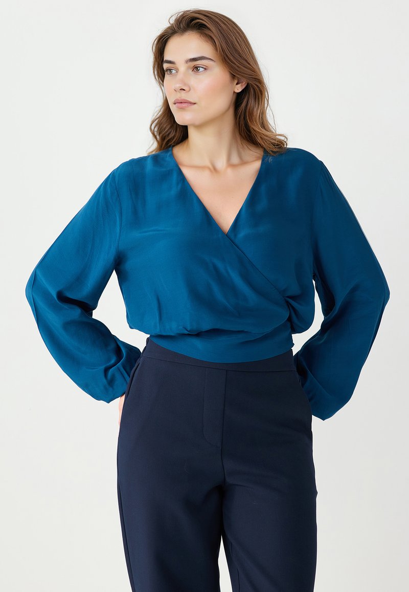 Blouse teal à manches longues avec un design enroulé, mettant en avant un décolleté en V profond et un tissu doux et lisse. Associée à un pantalon bleu marine foncé.