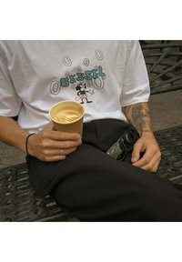 Camiseta gráfica blanca con un personaje de dibujos animados y el texto "BESSEL". Pantalones negros, sosteniendo un latte en un vaso de papel, con gafas de sol cerca.