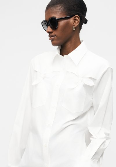 Femme portant des lunettes de soleil ovales noires et des boucles d'oreilles créoles dorées, vêtue d'une chemise blanche boutonnée avec des rabats de poche texturés, tournée vers la gauche.