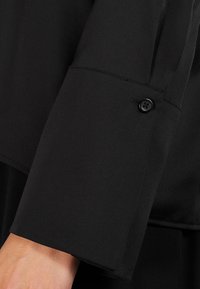 Blusa nera a maniche in tessuto liscio, con un bottone decorativo sul polsino. Il design include una cucitura pulita senza motivi.