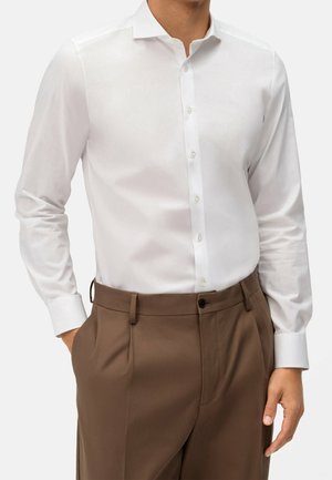 Mężczyzna ubrany w białą, długą koszulę zapinaną na guziki, włożoną w brązowe plisowane spodnie, z rękami częściowo w kieszeniach.