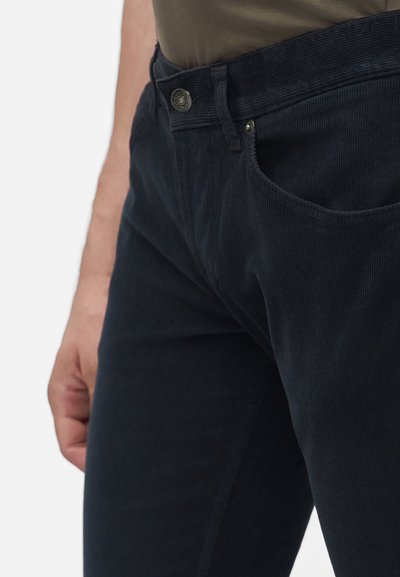 Gros plan d’une personne portant un pantalon en velours côtelé bleu foncé avec une poche avant visible, un bouton et une partie d’une chemise verte.