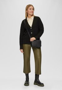 Schwarze Strickjacke mit Knöpfen, beiges kragenoberteil, olivgrüne Kunstlederhose und schwarze Ankle-Boots mit dicken Sohlen. Schwarze Umhängetasche.