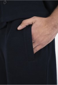 Main partiellement insérée dans la poche avant d'un pantalon noir texturé, montrant les détails du tissu et des coutures.