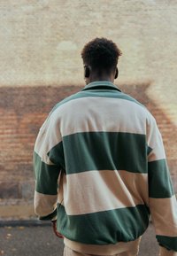 Personne aux cheveux courts et bouclés portant une veste rayée verte et beige, debout à l'extérieur face à un mur en briques.