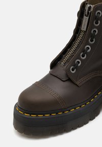 Bota de cuero marrón con punta redonda, cierre de cremallera, ojales de metal y costuras amarillas sobre una gruesa suela de plataforma de goma negra.