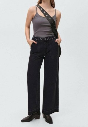 Donna con canotta grigia, jeans neri a gamba larga con cintura con occhielli, borsa a tracolla nera e stivaletti neri, mani nelle tasche.