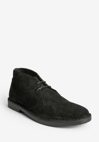 Botín chukka de ante negro con puntera redondeada, cordones de cuero y suela plana de goma. Presenta costuras a lo largo de la suela y un diseño minimalista.