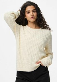 Crème kabelbrei sweater met een ronde halslijn en lange mouwen, met een gestructureerd patroon en geribbelde manchetten, gecombineerd met donkere broeken.