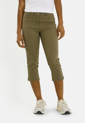 Personne portant un pantalon court vert olive, des baskets blanches, un haut blanc et un bracelet doré, debout sur un fond blanc.