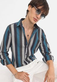 Striped long-sleeve shirt in blue, gray, and navy, made of a soft fabric. Features a buttoned front and a relaxed collar. White pants visible.