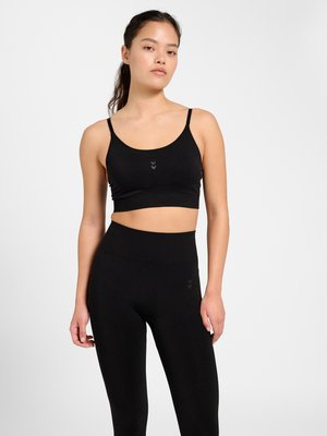 Ung kvinde iført sort sports-BH og leggings med høj talje står mod en ensfarvet hvid baggrund med afslappet kropsholdning og neutralt ansigtsudtryk.