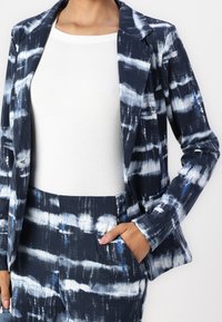 Blazer bleu marine avec motif abstrait blanc et bleu, fermeture à un bouton, et revers crantés, associé à un top blanc côtelé.