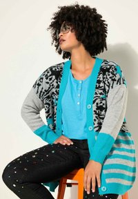 Cardigan con motivi neri e grigi, dettagli turchesi e chiusure a pulsante, abbinato a un top azzurro chiaro e jeans neri con gemme.