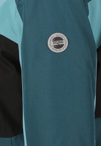Türkisfarbener und schwarzer Sportjackett mit einem runden silbernen Logo-Patch, auf dem "ZIGZAG" steht. Hergestellt aus einem glatten, strukturierten Stoff; zeichnet sich durch ein kontrastierendes Design der Paneele aus.
