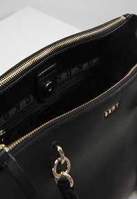 Svart läderhandväska med en texturerad yta, guldfärgad dragkedja och metallhaltare. Insidan har ett märkesinklädnad och ett tryckknappstängning.