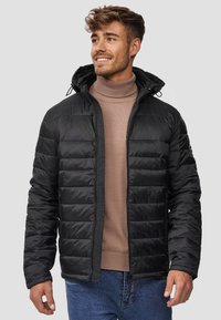 Doudoune noire avec fermeture éclair à l'avant, poignets élastiques et capuche, présentant un design matelassé. Portée par-dessus un pull en col roulé marron clair.