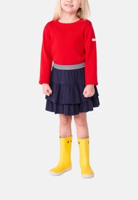 Maglione rosso lavorato a maglia, gonna a balze blu navy con cintura a righe, e stivali di gomma gialli con dettagli intagliati.