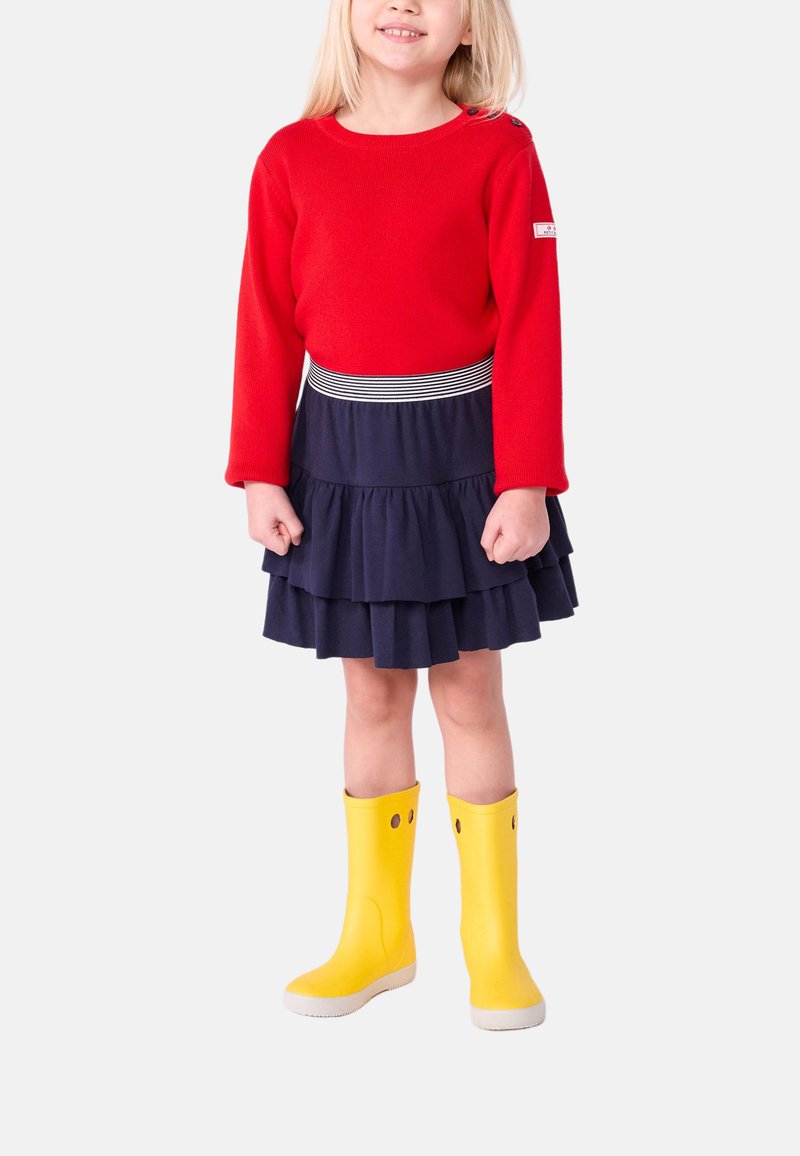 Maglione rosso lavorato a maglia, gonna a balze blu navy con cintura a righe, e stivali di gomma gialli con dettagli intagliati.