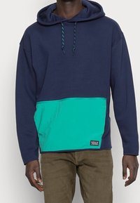 Man som bär en marinblå huvtröja med en teal-färgad ficka och snören i fram, i kombination med bruna byxor, står mot en enkel bakgrund.