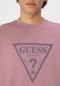 Rózsaszín pulcsi kerek nyakkivágással és bordázott díszítéssel. Sötétszürke háromszöglogóval rendelkezik, amelyen a "GUESS ORIGINALS" és a "WASHED JEANS" felirat szerepel.