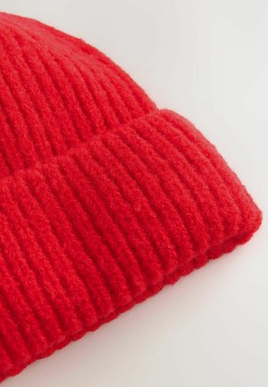 Nahaufnahme einer strukturierten roten Beanie, die über gerippte Details und einen umgeschlagenen Rand verfügt, vor einem hellen Hintergrund.