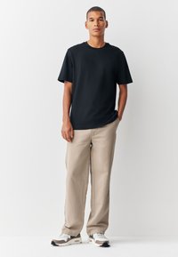 Juoda marškinėliai su apvalia kaklo iškirpte ir trumpomis rankovėmis, derinami su šviesiai smėlio spalvos kelnėmis ir baltais sportbačiais su rudais akcentais. Paprasta, kasdienė apranga.