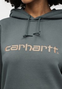 Σκούφο με κουκούλα σκούρου πράσινου χρώματος με μεγάλο κεντημένο λογότυπο "carhartt" σε μπεζ χρώμα. Ρυθμιζόμενα κορδόνια στη κουκούλα με υφή.